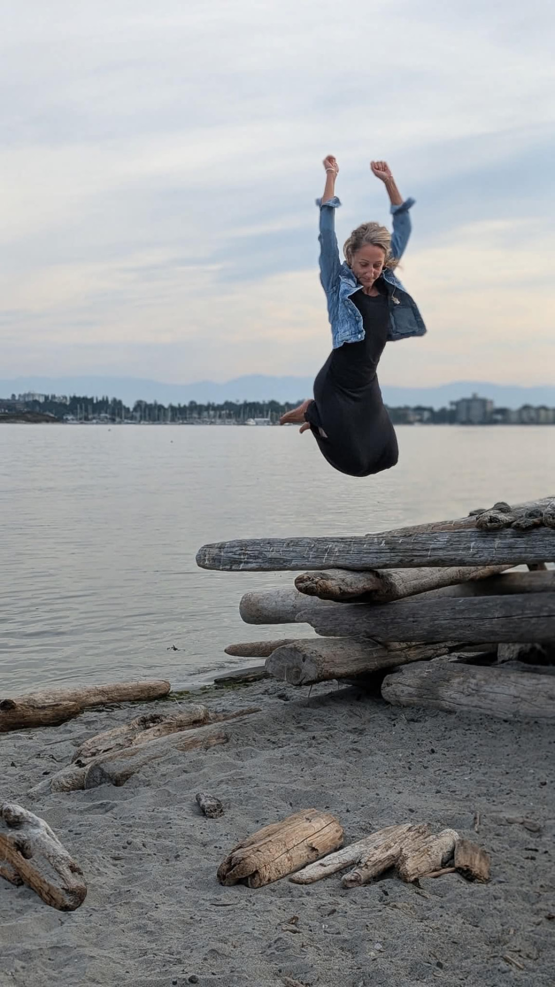 Paula Kelene leaping with arms raised on a driftwood beach at dusk